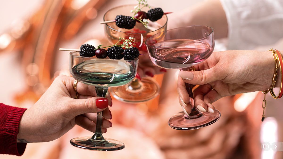 Das Bild zeigt drei Hände, die elegante Cocktailschalen anstoßen. Die Gläser sind mit dekorativen Beeren garniert, darunter Brombeeren und Kirschen, und haben unterschiedliche Farben: rosa, grün und bernsteinfarben. Die Szene vermittelt ein Gefühl von Geselligkeit und Feierlichkeit.