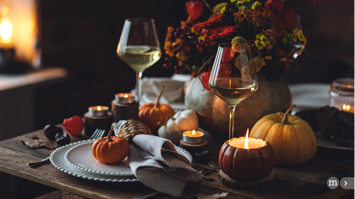 Ein gemütlich gedeckter Herbsttisch mit einer brennenden Kerze, die in einen kleinen Kürbis geschnitzt ist. Auf dem Tisch stehen mehrere unbeleuchtete Kerzen und zwei Gläser Weißwein. Ein kunstvoller Strauß aus Herbstblumen und -blättern in warmen Farben, zwei kleine Kürbisse und Tannenzapfen vervollständigen das Arrangement. Die Atmosphäre ist intim und warm, was auf einen festlichen oder feierlichen Anlass im Herbst hinweist.